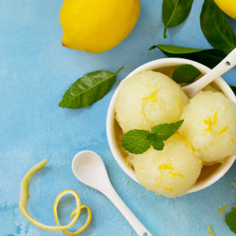 3 scoops of Lemon Sorbet, made with Special Ingredients Xanthan Gum, set in a delicate bowl, surrounded by green leaves and half lemons.